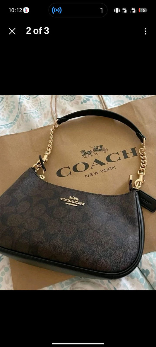 A Coach shoulder bag in the signature dark brown monogram canvas with gold-tone chain and strap hardware, resting on a Coach New York paper shopping bag.