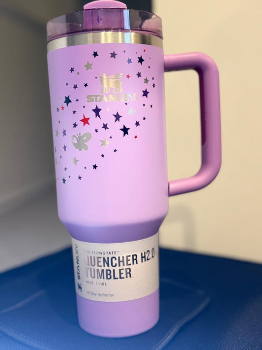 A lavender purple Stanley tumbler with a handle, decorated with silver, red, and blue stars, and a white label near the bottom.