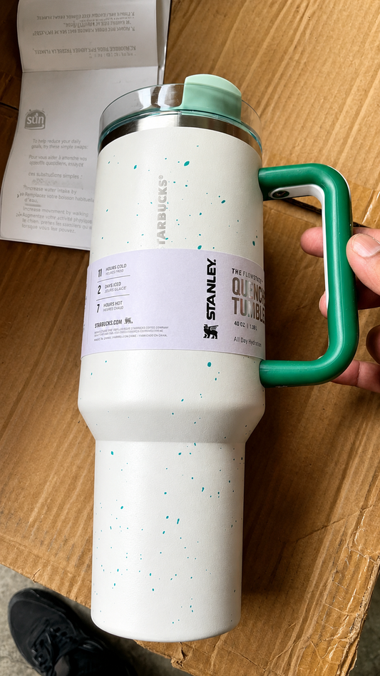 A white Starbucks Stanley 32 oz insulated tumbler with teal speckles, a green handle, and a clear lid, held by a hand on a cardboard box.