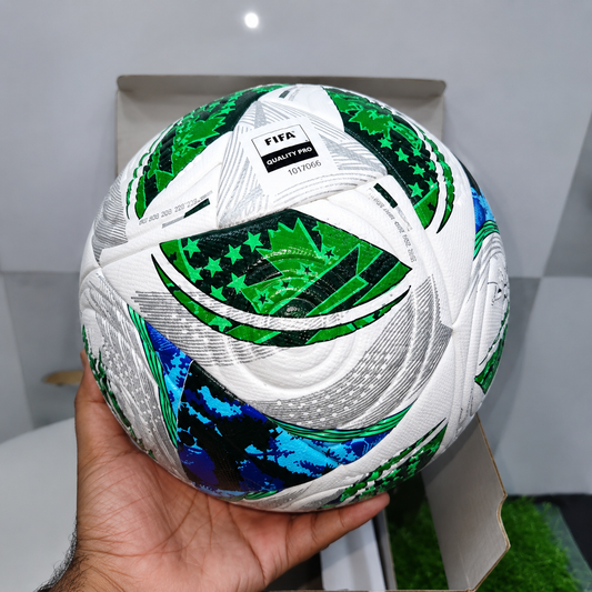 A hand holding a white FIFA 2026 World Cup soccer ball with green, blue, and black patterned panels, positioned in front of an open cardboard box.