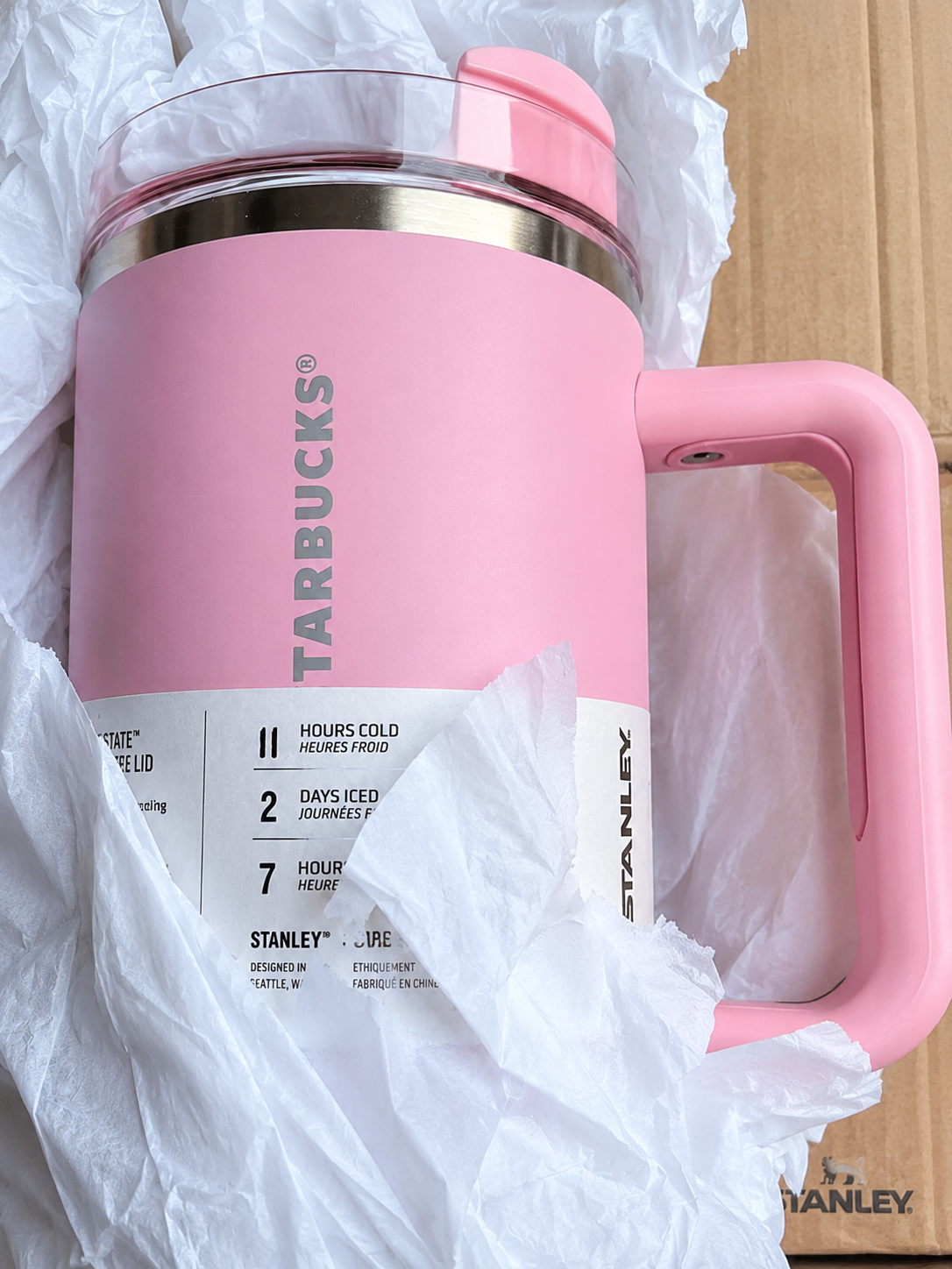 A pink Starbucks travel mug with a handle, shown wrapped in white crumpled paper and cardboard packaging. The mug features a metallic rim, a clear lid, and a white label detailing product features.