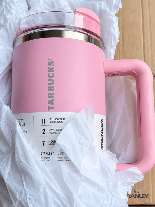 A pink Starbucks travel mug with a handle, shown wrapped in white crumpled paper and cardboard packaging. The mug features a metallic rim, a clear lid, and a white label detailing product features.