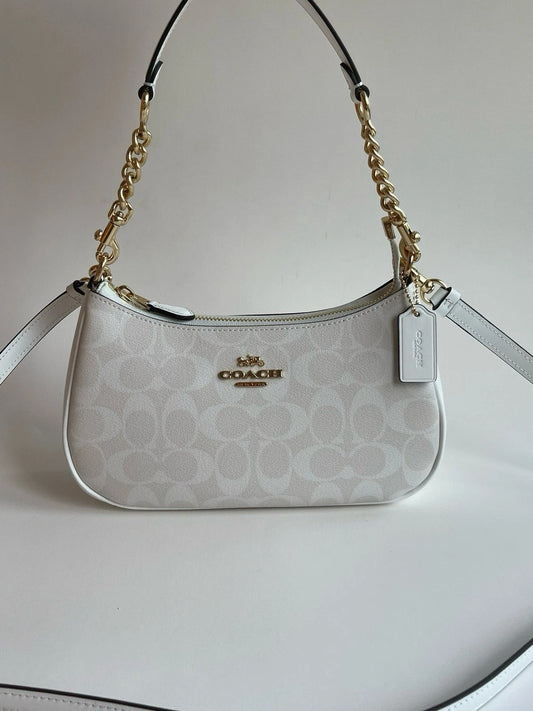 A Coach shoulder bag in white and beige signature monogram canvas with gold-tone hardware, a top zipper, and a hanging brand tag, photographed against a plain light background.