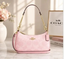 A light pink Coach shoulder bag with a gold-tone chain strap and a gold logo plate, displayed on a light-colored surface next to a vase of flowers and a framed Coach logo.